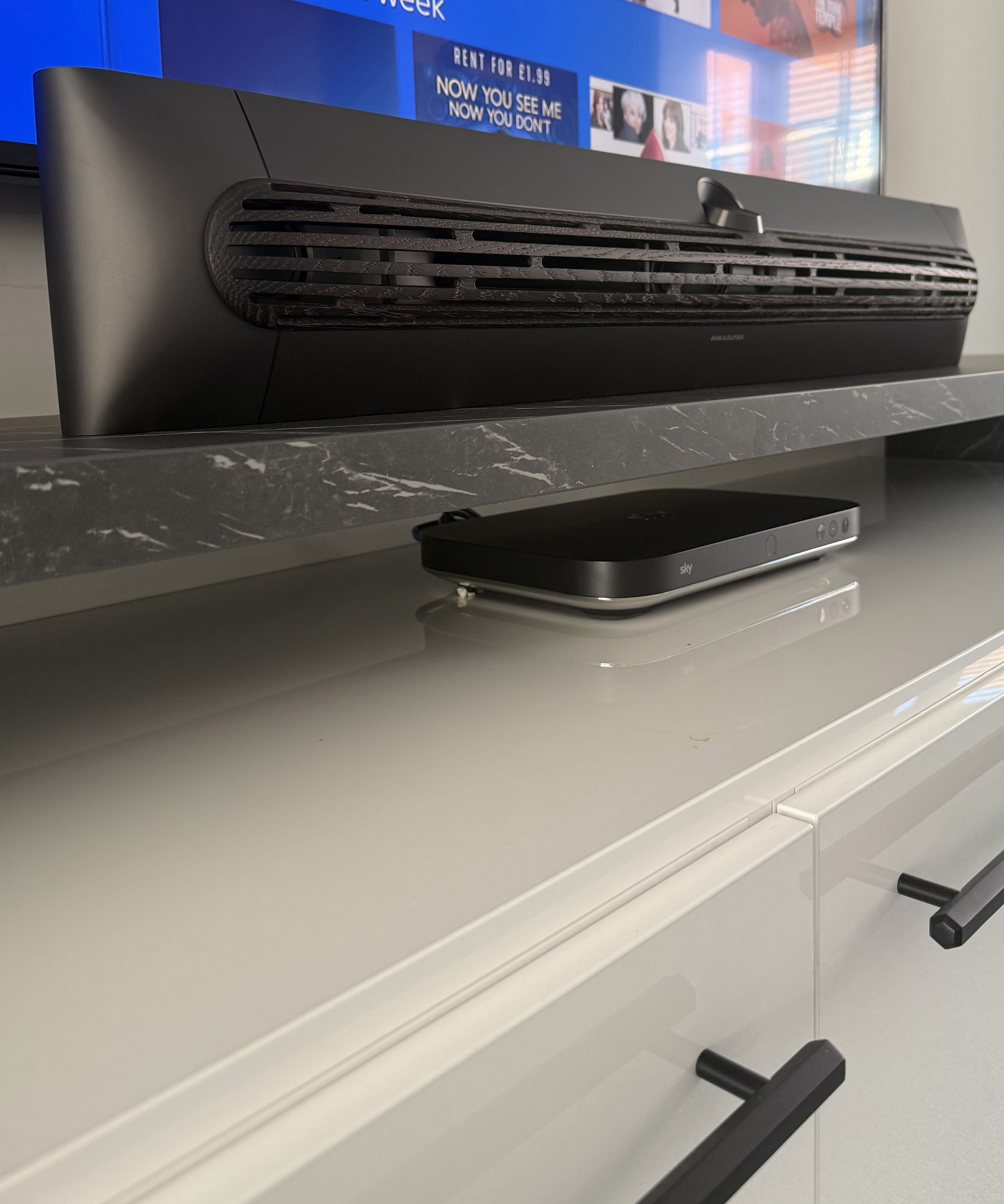 Angled close-up of the soundbar on a TV unit, highlighting its textured front grille and matte finish