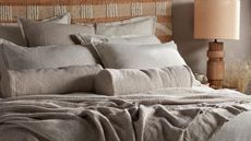 Greige linen bedding with piles of throw pillows, a wooden bedside lamp with shade beside it
