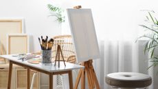 A bright white craft room with an art easile and blank canvas. A table with paints and brushes beside it 