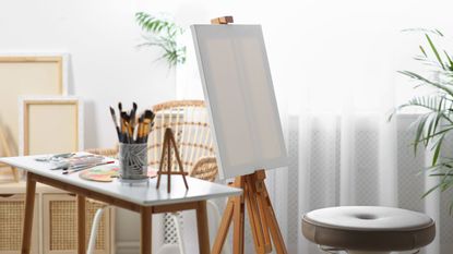 A bright white craft room with an art easile and blank canvas. A table with paints and brushes beside it 