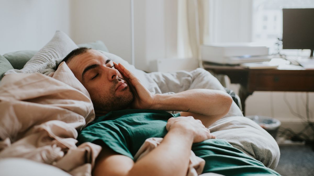 A sleep deprived man in a green shirt falls asleep for a second on his sofa, having what experts refer to as a microsleep because he&#039;s sleep deprived