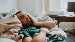 A sleep deprived man in a green shirt falls asleep for a second on his sofa, having what experts refer to as a microsleep because he's sleep deprived
