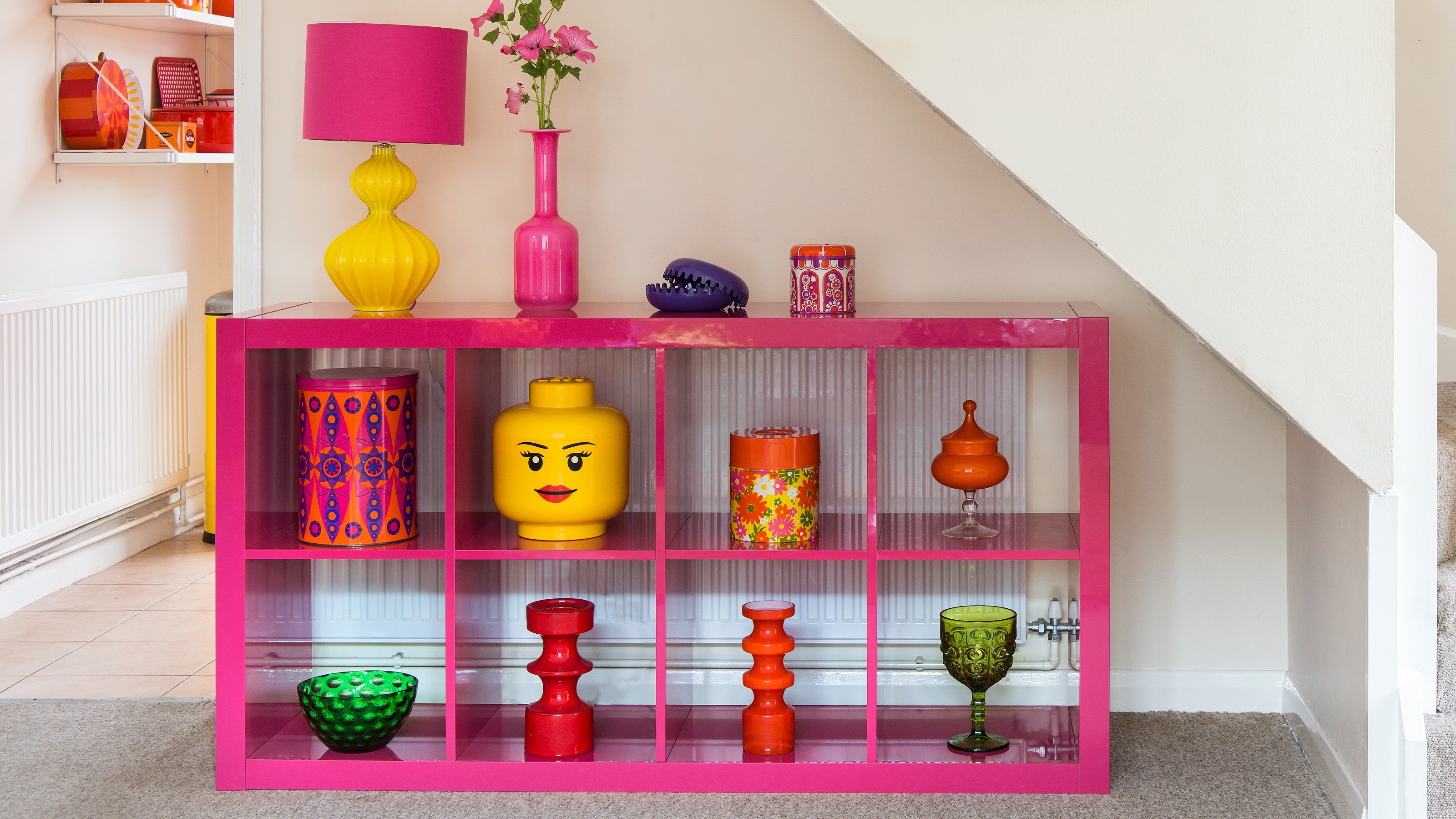 A hallway with a pink glossy IKEA KALLAX storage unit placed under the stairs displaying colourful decor