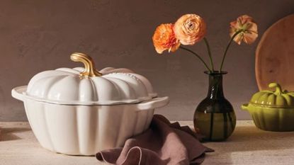 A white Le Creuset Pumpkin cocotte on a counter beside a black glass vase of orange flowers.