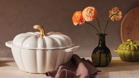 A white Le Creuset Pumpkin cocotte on a counter beside a black glass vase of orange flowers. 