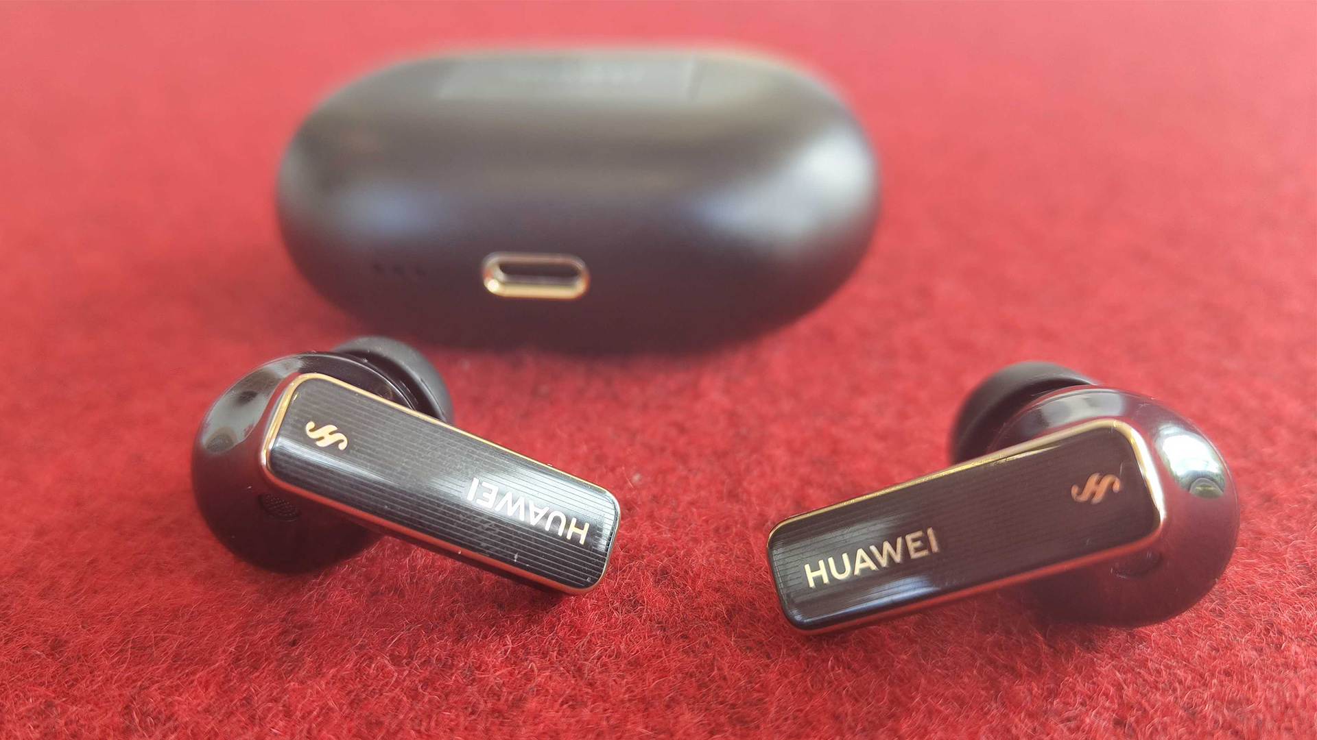 Huawei FreeBuds Pro 4 in-ear headphones on red fabric surface in front of charging case