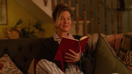 Image of actress Renée Zellweger as Bridget Jones wearing striped pajamas and a dark cardigan. She is sitting on a dark green sofa with patterned throw pillows and throw blankets around her. She is holding a red diary.