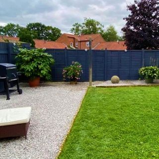 Potted plants on gravel next to lawn, barbecue and wooden blue fence.