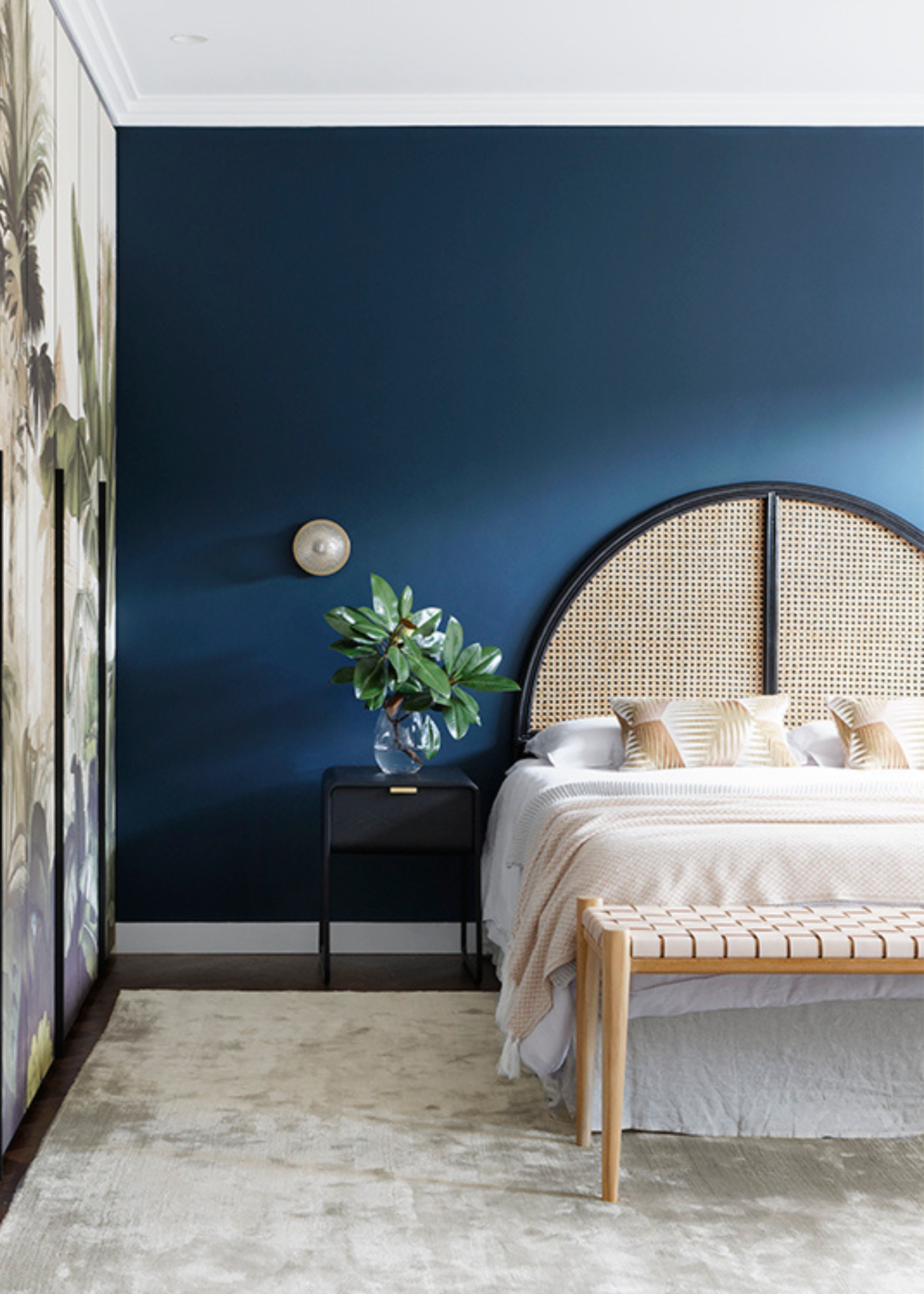 A blue bedroom with a white bed, a gray rug, a black side table, a tan and white bench, and a rattan headboard