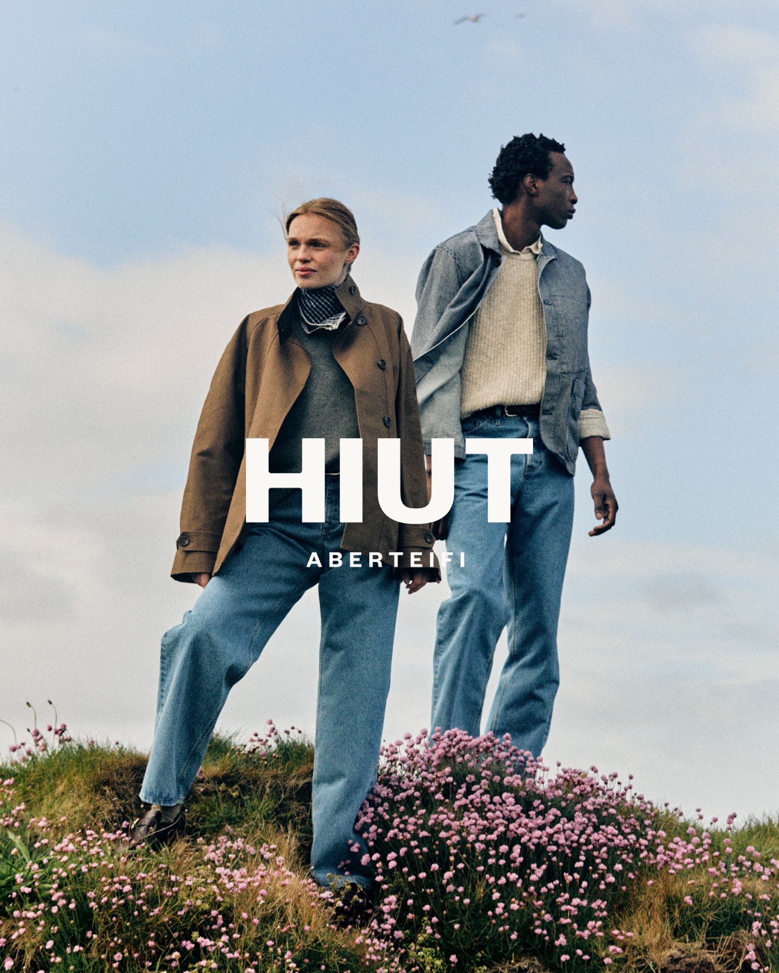 Two models stand outdoors on a grassy hill covered in pink wildflowers under a pale blue sky, with the brand name "HIUT ABERTEIFI" overlaid in bold white text across the center.