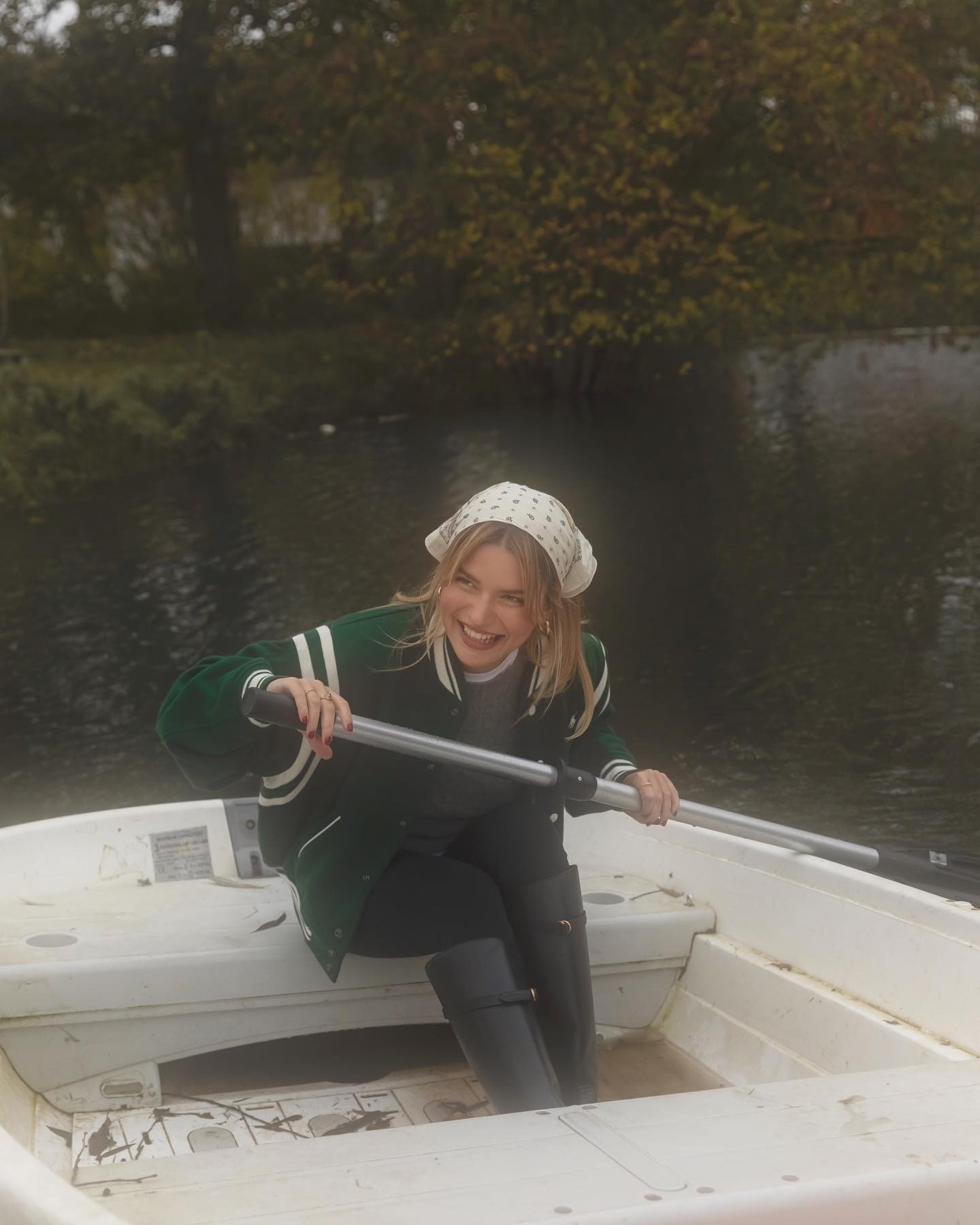 Fashion person Sabina Socol wears a leggings outfit 2025. She wears a green baseball jacket, black leggings, grey jumper and riding boots.