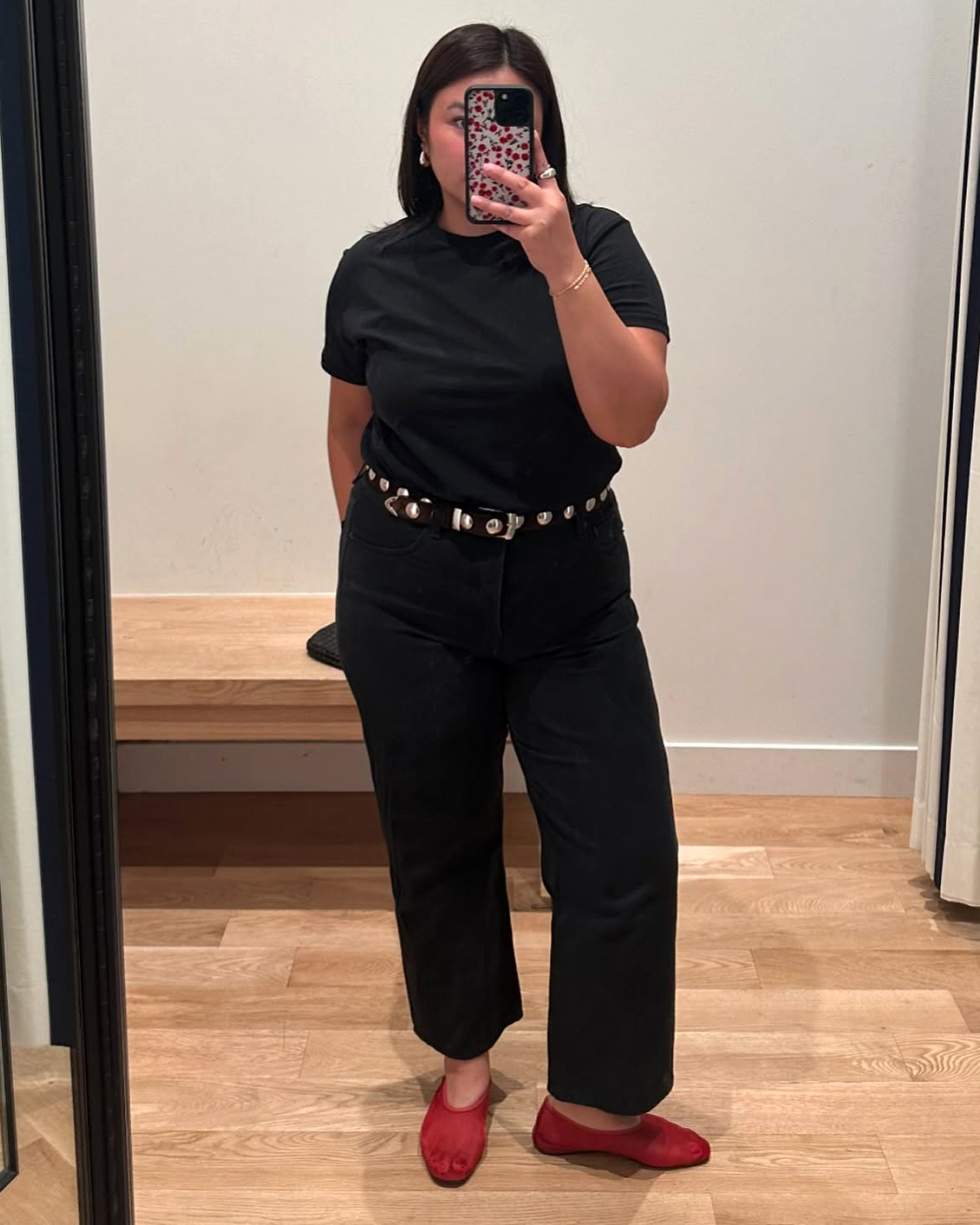 Style influencer Marina Torres poses for a dressing room mirror selfie wearing a black T-shirt, a studded belt, cropped black jeans, and red mesh ballet flats.