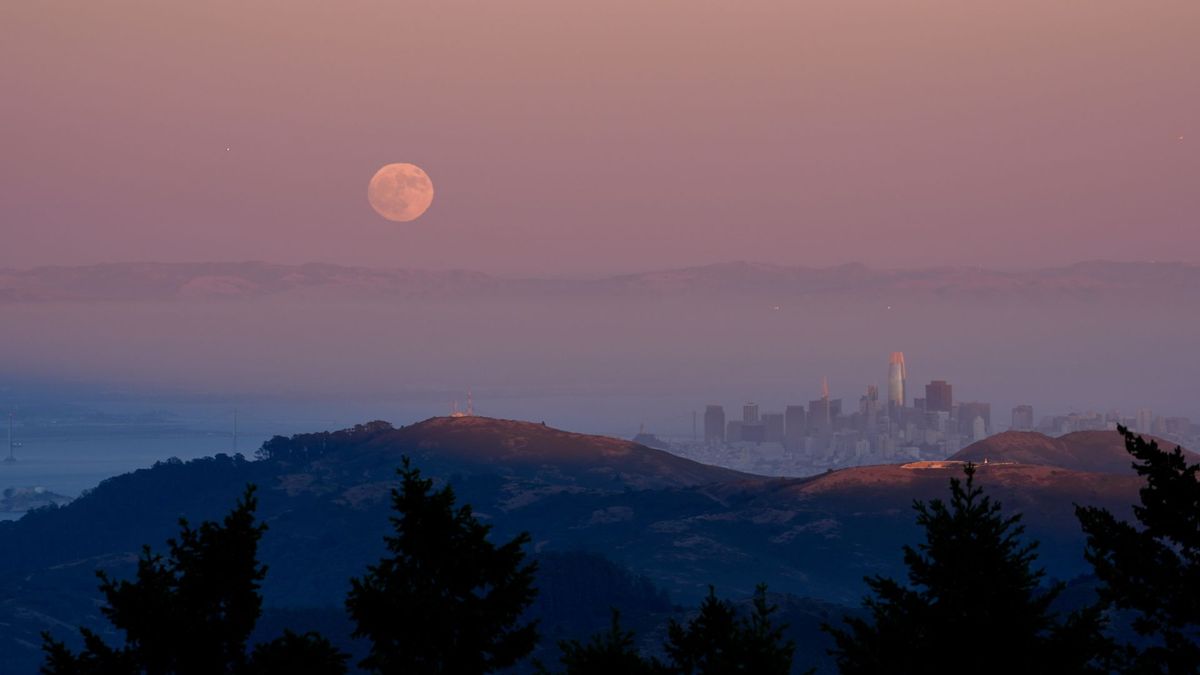 La luna llena se eleva en un cielo color melocotón sobre las colinas cercanas a una ciudad, cuyos rascacielos se pueden ver a la derecha de la foto.