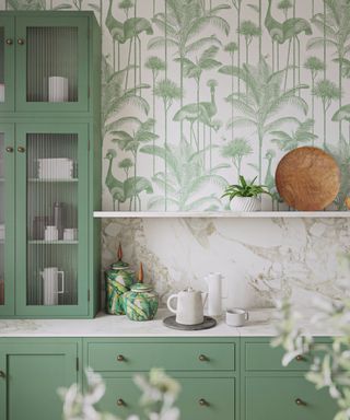 A kitchen with green kitchen cabinets and shelves, green and white leafy wallpaper, and white shelves and countertops