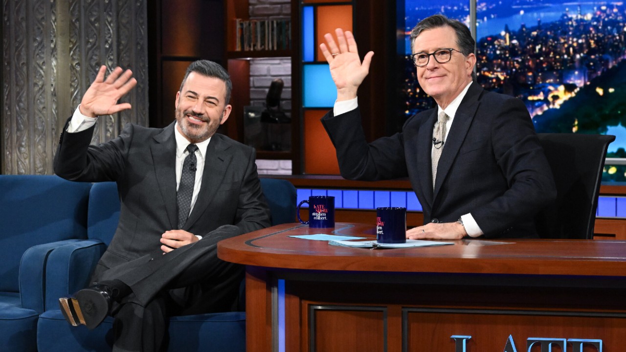 From left to right: Jimmy Kimmel and Stephen Colbert sitting next to each other on The Late Show, they're both waving with their left hands. 
