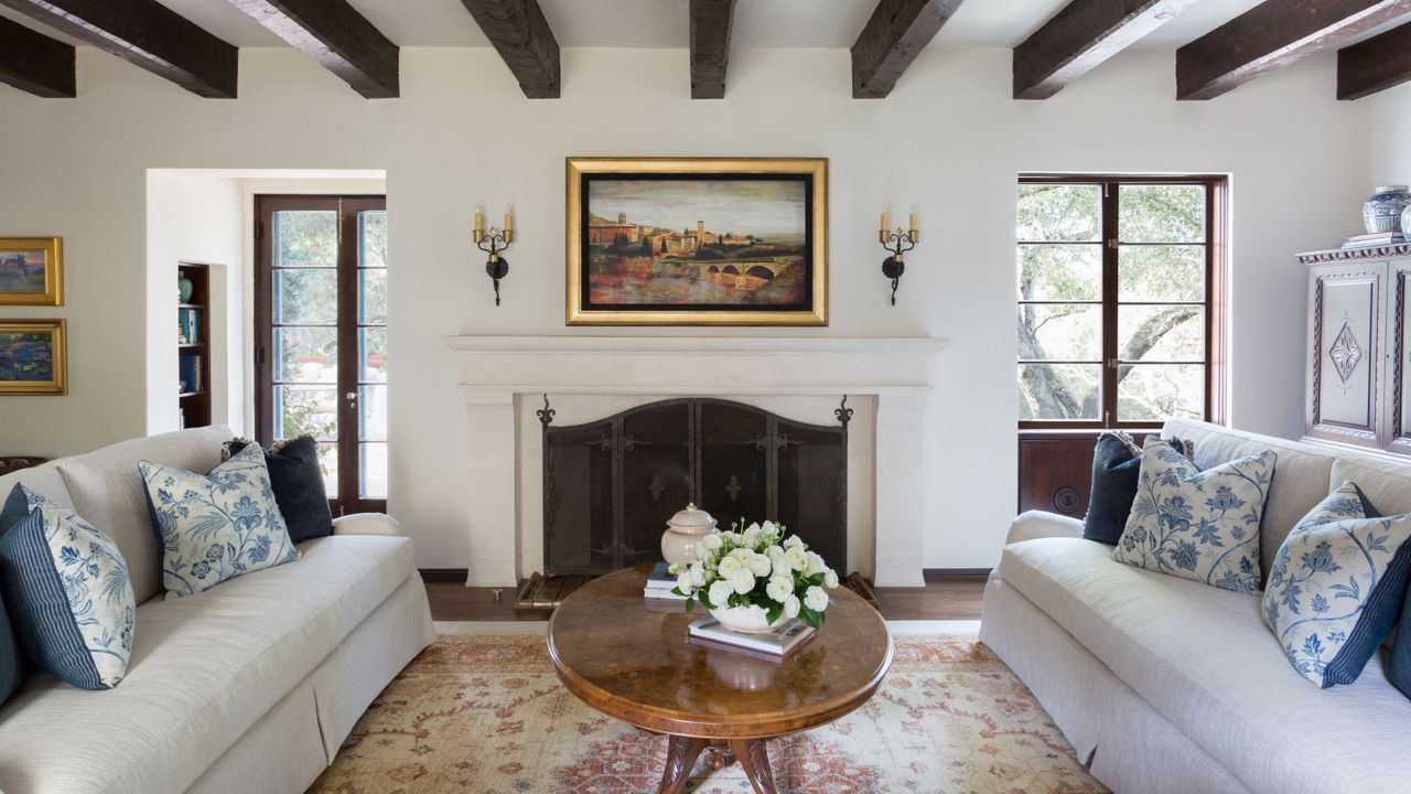 traditional living room with dark ceiling beams and white sofas and stone fireplace with gilt framed picture above and oval mahogany coffee table with oriental style rug