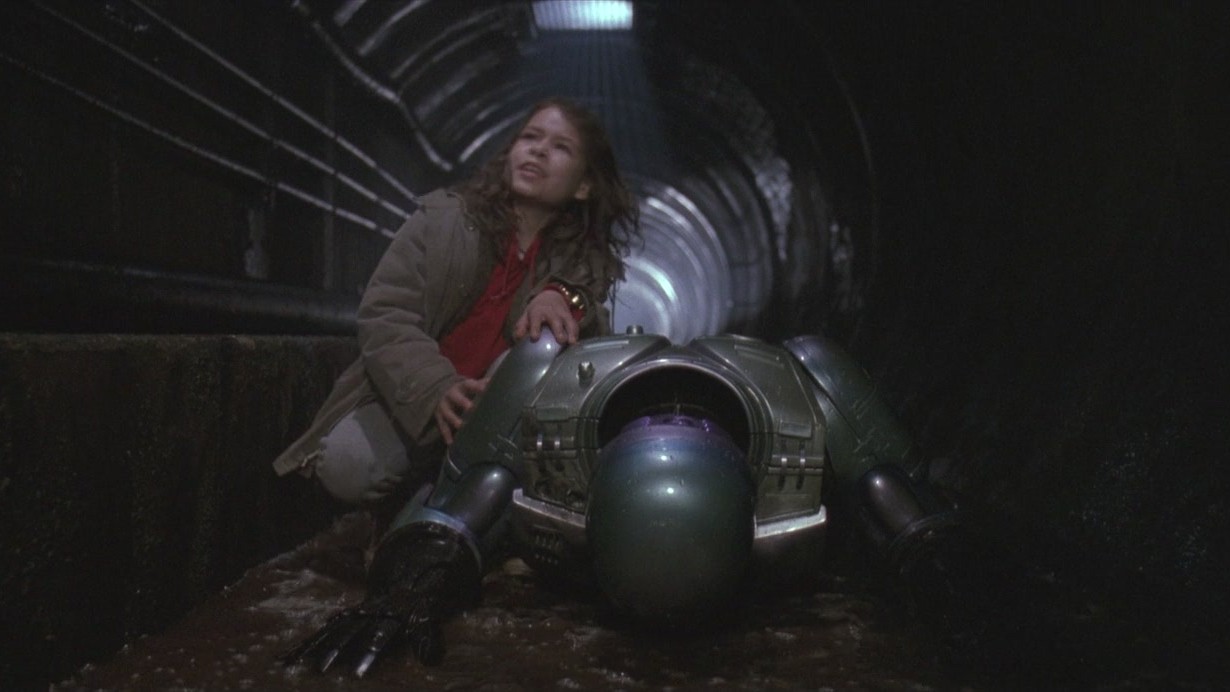 Still from the movie RoboCop 3 (1993).  Here we see RoboCop face down in a sewer with his young sidekick trying to help him up.