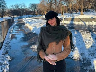 A photo of Kendall Jenner wrapping up in an olive green fringe shawl, a brown suede jacket, and a black newsboy cap for a walk.