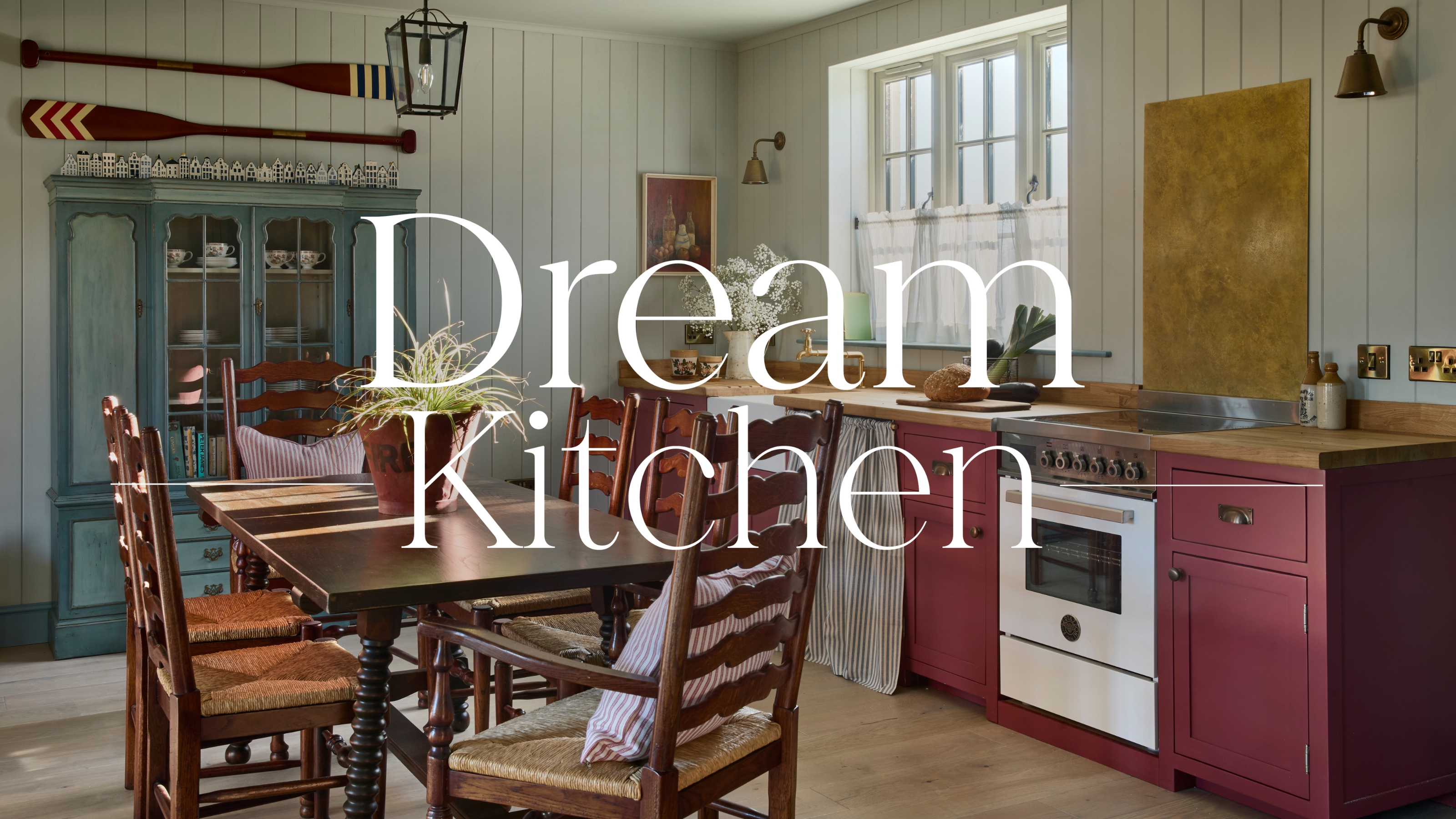 A Scottish coastal cottage kitchen with soft blue paneled walls, burgundy cabinets, a wooden kitchen table, and a dark blue antique cabinet