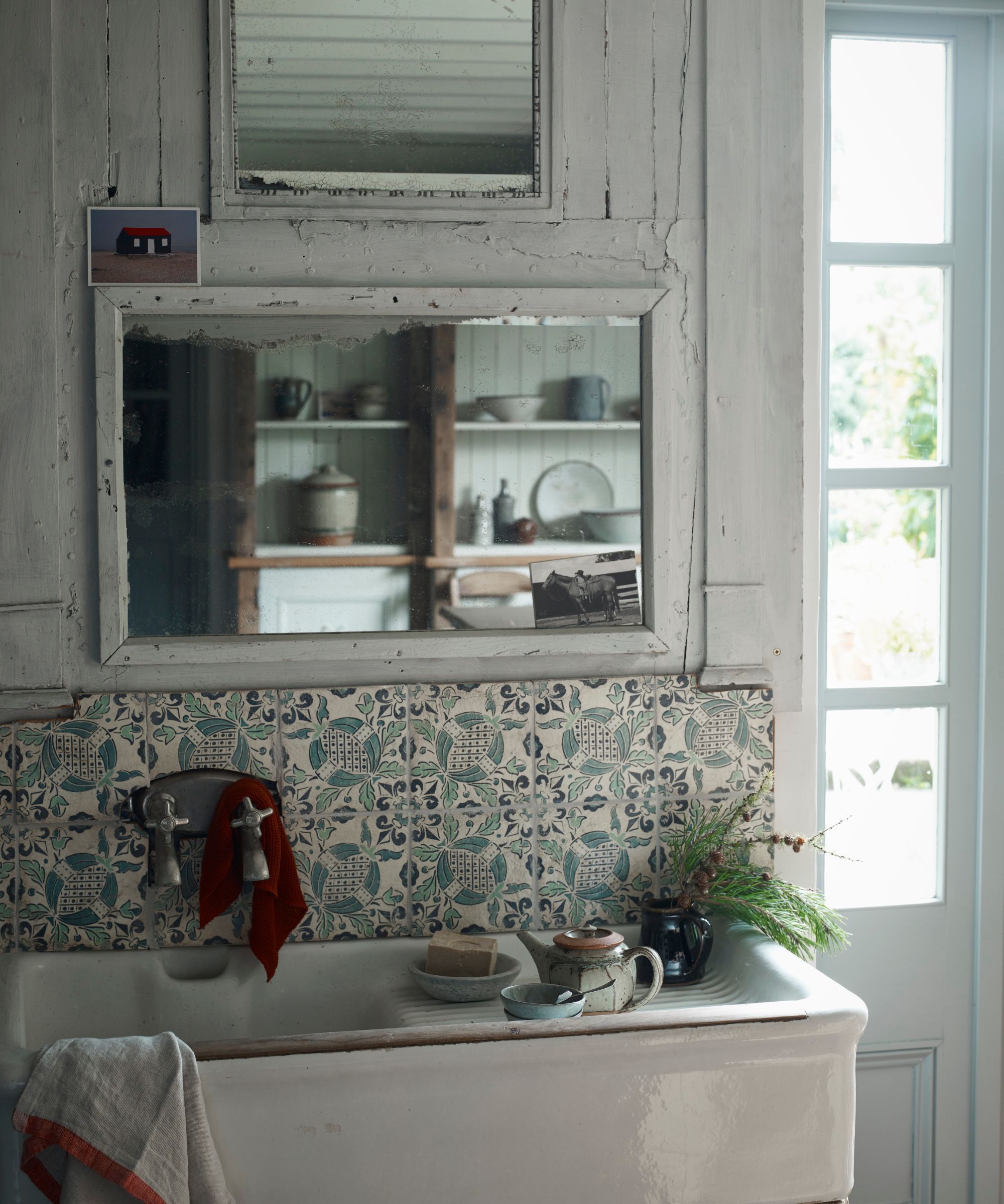 A white trough sink with a blue patterned backsplash, and a two mirrors above. The walls are white, and to the right is a panelled window panel.