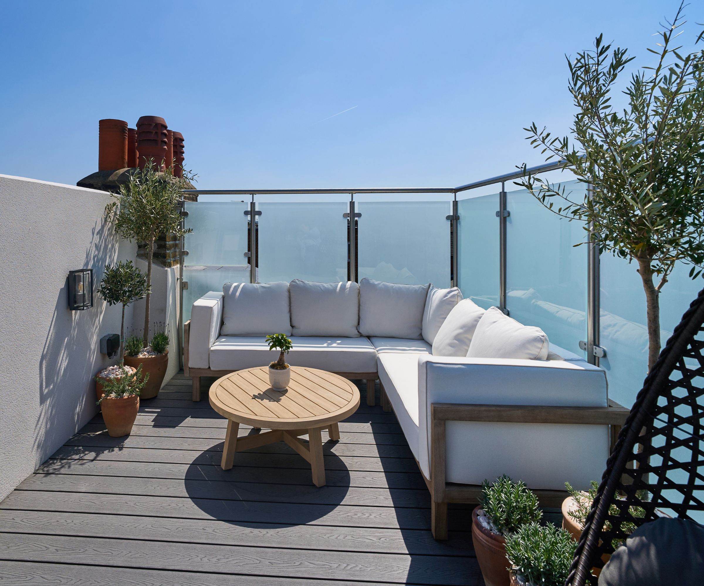 Outdoor rooftop terrace with L-shaped white cushioned sofa, potted plants and round wooden coffee table, enclosed by frosted glass balustrades.