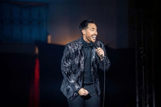 Colin (Ross Adams) stands on stage in the ship's theatre, wearing a black shirt, a black bow tie, and a black blazer with sequins, holding a microphone and talking to the crowd