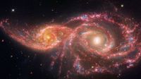 adjacent spiral galaxies with glowing arms of reds and pinks stare like a pair of eyes, their centers white hot yellow, from the black abyss of space.