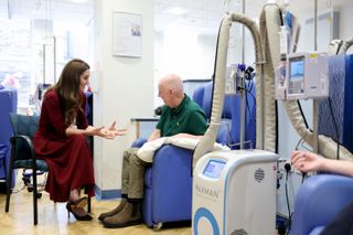 Kate Middleton wearing a red sweater and skirt talking to a man receiving cancer treatment in a blue chair at a hospital