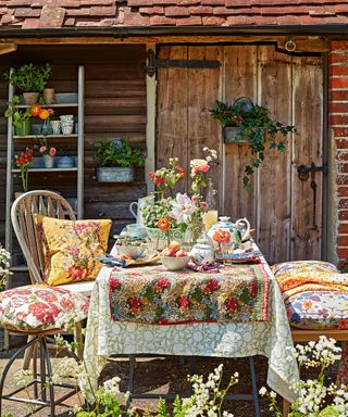 A brightly colored table setting with floral tablecloth and cushions in front of a garden shed.