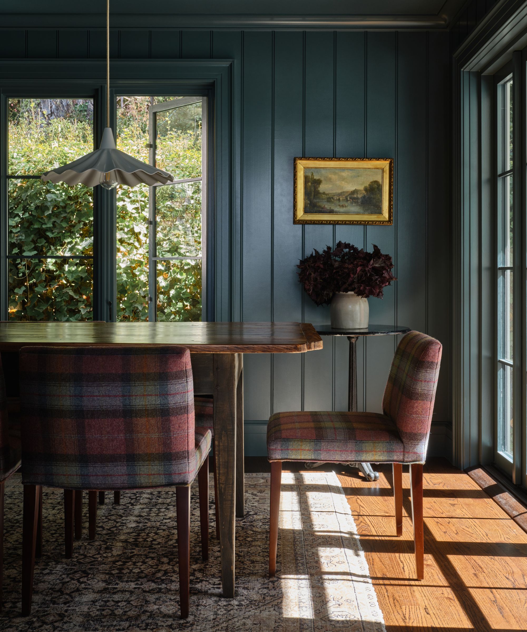a cozy dining room with plaid upholstered dining chairs, a vintage-inspired persian rug on the floor, a large wood table, blue-painted wood panels on the walls, and a pendant hanging over the table