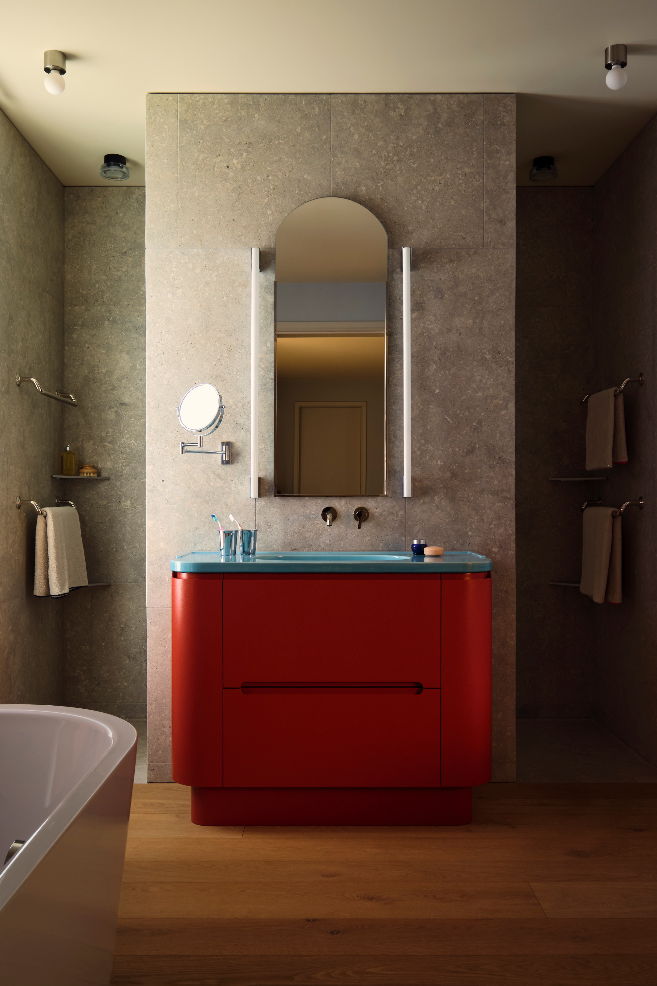 Bathroom with grey stone walls, wood floor, red vanity with blue countertop, curved mirror