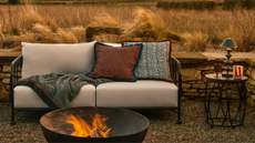 an outdoor sofa with cushions and a throw blanket with a firepit in front, and a side table with a lit portable table lamp, glass and candle