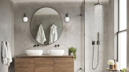 A neutral bathroom with an open shower with a glass partition next to a wooden wall-mounted bathroom vanity with dual sinks. A large round mirror mounted on the wall above them. 