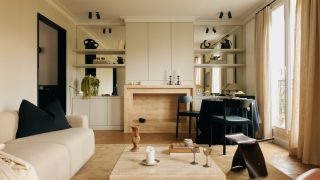 Living room with a cream sofa, travertine coffee table, black dining chairs, mirrored wall shelving, beige curtains and herringbone wood flooring.