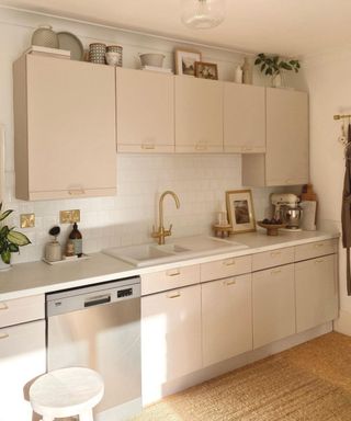 A stylish neutral modern small kitchen with brass hardware, warm neutral cabinets, white wall tiles, and kitchenalia stored on top of cupboards