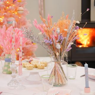 christmas flower arrangement in tones of pink peach and purple on a table setting of the same theme with candles and mince pies and a fireplace burning in the background
