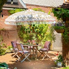 A garden with patio with a wooden dining table and chairs and a decorative parasol surrounded by planting and flowers with hanging baskets