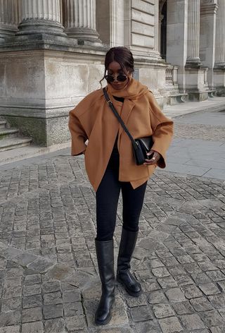 A winter travel outfit is shown in a photo of a woman standing outside wearing a tan scarf coat over a black sweater with black leggings, black riding boots, a black crossbody bag, and black sunglasses