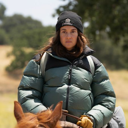 A woman (Shailene Woodley as Annie Clay) wears a black "Graceland" beanie and a puffy green jacket while riding atop a horse outdoors, in 'Paradise' season 2.