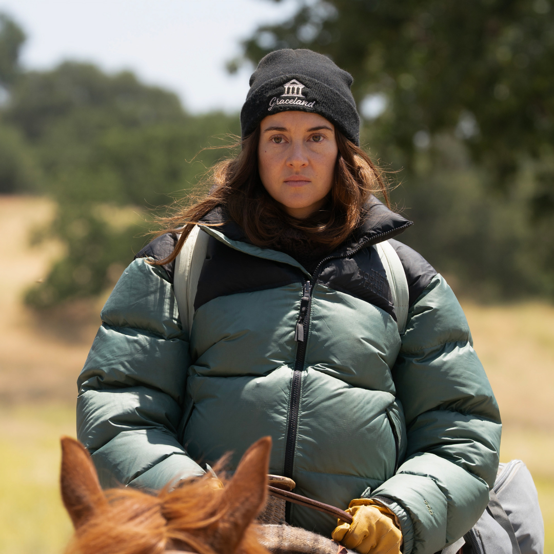 A woman (Shailene Woodley as Annie Clay) wears a black "Graceland" beanie and a puffy green jacket while riding atop a horse outdoors, in 'Paradise' season 2.