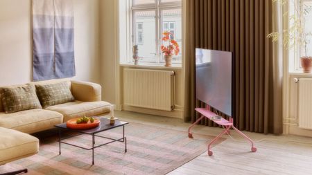 Pedestal pink Moon Rollin' tv stand in living room with beige L-shape sofa, pink and gray stripe rugs, coffee table, wall hanging, and beige curtains