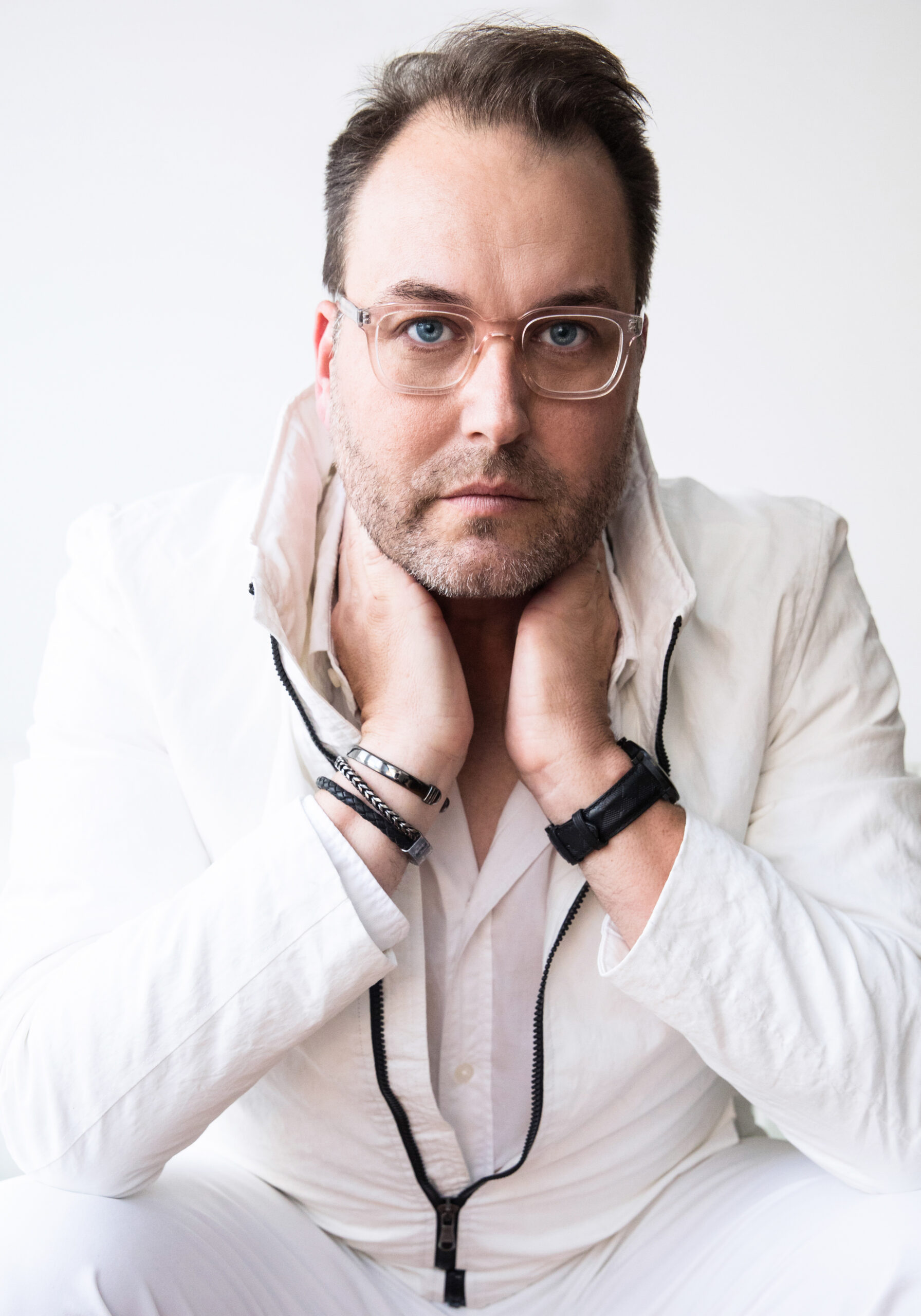 A man with square, transparent glasses, dressed in an-all white casual-chic outfit with black armbands and jewelry poses for the camera while sitting with his hands on his neck.