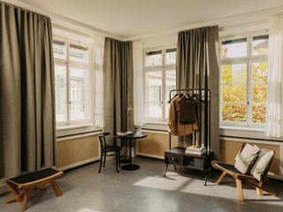 A room in the hotel. Dark grey floors dark wooden chairs and a table sit in the corner. A clothes rack stands next to the wall. Grey curtains are open revealing large windows.