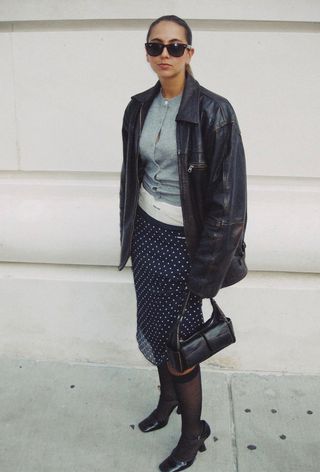 A woman wearing a leather jacket over a gray cardigan with exposed white boxer Miu Miu briefs, a polka dot sheer skirt, black sheer knee-high socks, and ankle strap pumps.