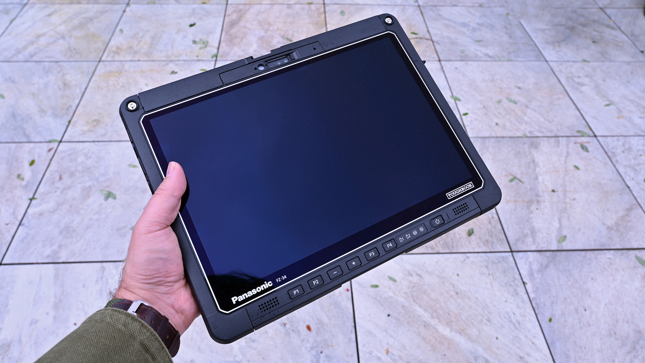 A hand holding a Panasonic TOUGHBOOK FZ-34 tablet outdoors, showing its black screen and front control buttons.