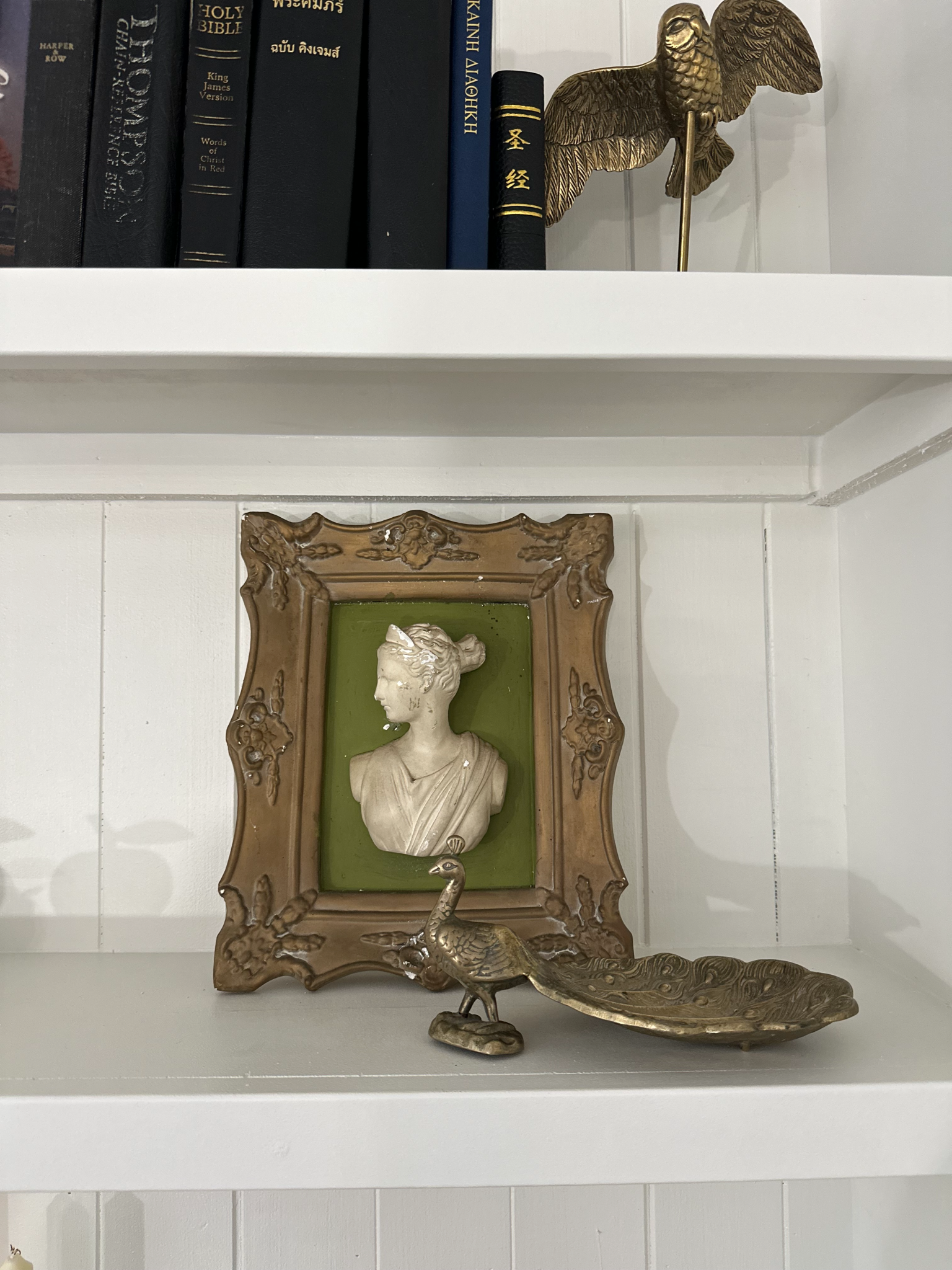 Close up of white shelves displaying a row of books, a photo frame with a bust silhouette, and brass figurines