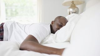 A man wearing a white t-shirt lies on his stomach on a bed, smiling because he's enjoying a good nap