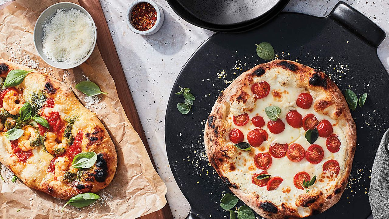 Two pizzas with one presented on a pizza baking stone and the other on a wooden chopping board with brown-colored baking paper