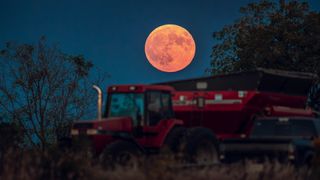A large full moon with a red/pink hue hangs low in the sky above farming machinery in the foreground.