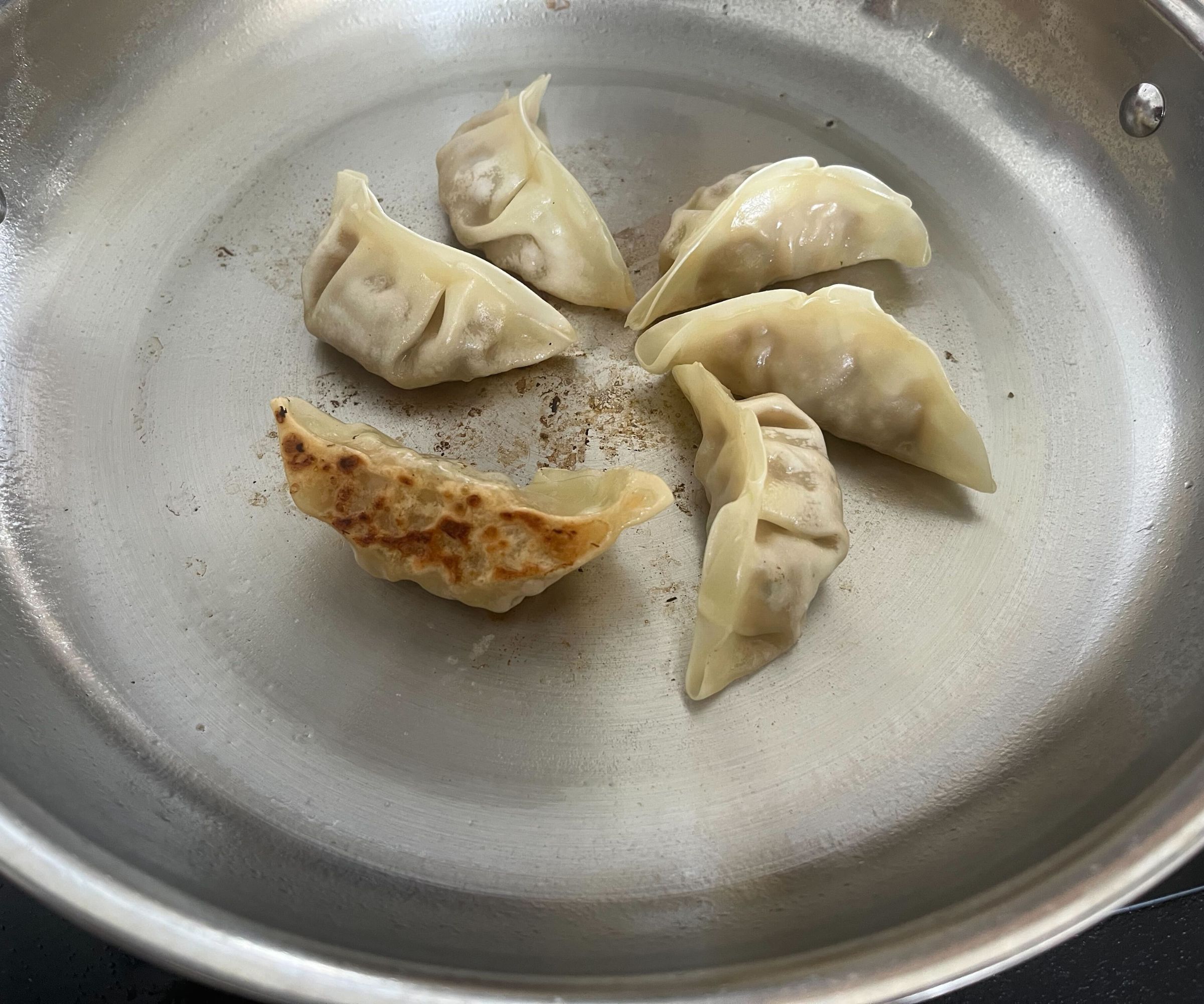 Gyoza dumplings being fried in a stainless steel frying pan, one on its side to show a crispy base.